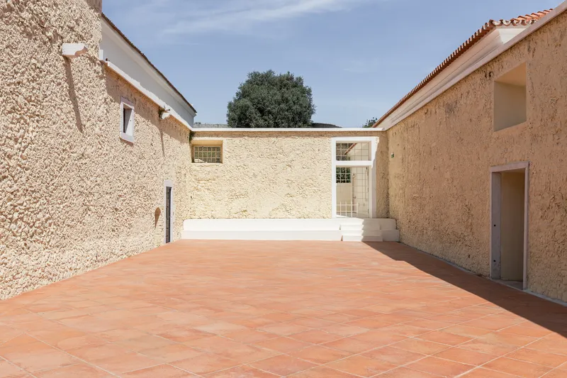Courtyard bounded by textured stone walls, terracotta paving, and a single tree framed by rooflines