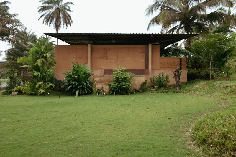 Rammed earth wall with timber shutters beneath a corrugated metal canopy surrounded by palms and lawn
