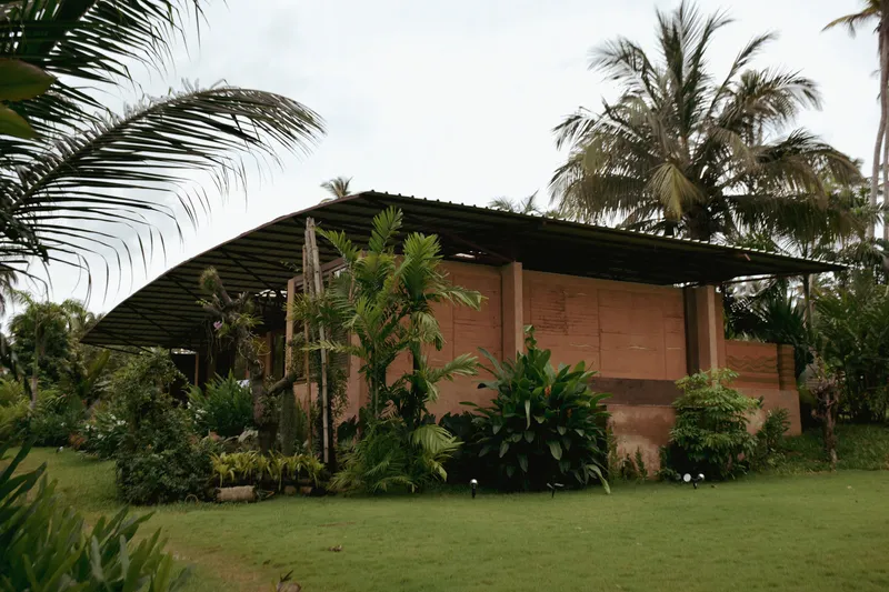 Rammed earth wall volume with wide corrugated roof overhang surrounded by palm trees and lawn