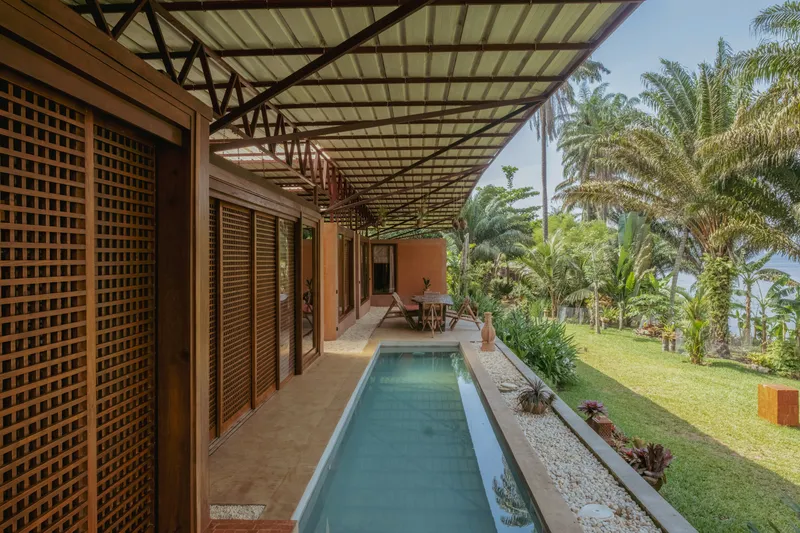 Lap pool alongside lattice-screened veranda with steel truss roof overlooking sloped lawn and palms