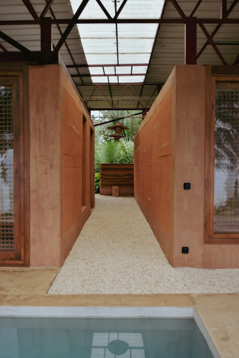 Gravel corridor flanked by rammed earth walls with exposed steel truss roof above a pool edge