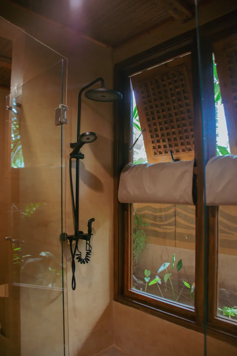 Glass-enclosed shower with black rainfall fixture and latticed wooden window opening to greenery