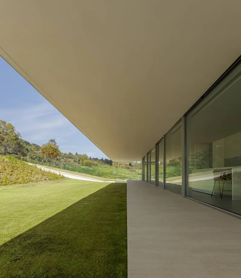Covered terrace with continuous glazing running parallel to a lawn sloping up the hillside