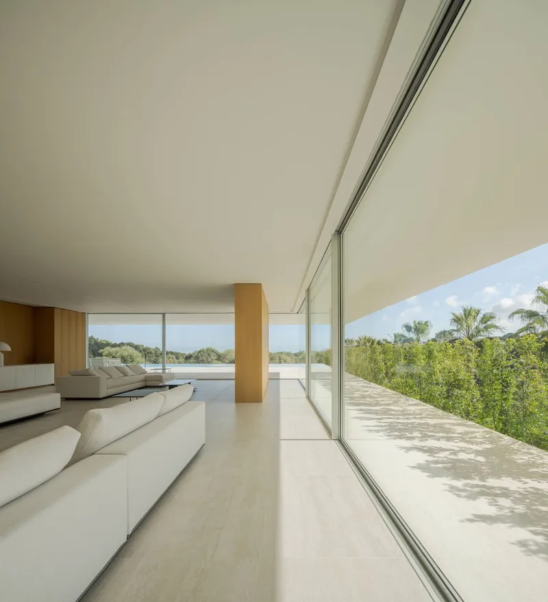 Living room with full-height glazing opening to a sun-dappled terrace and lush vegetation beyond