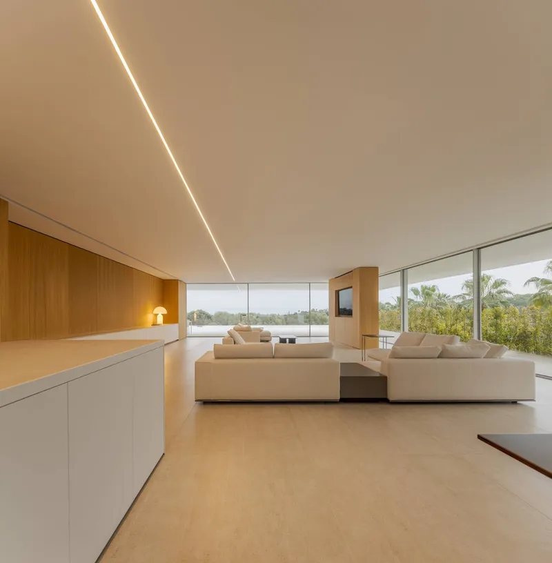 Open living room with continuous ceiling light slot and floor-to-ceiling glazing overlooking tropical landscape