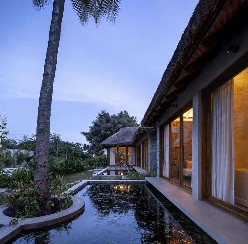 Reflecting pool extending between thatched roof volumes at dusk with palm trees overhead