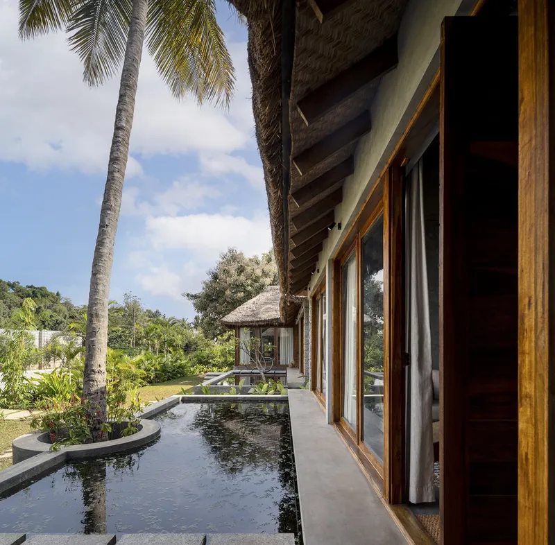Covered veranda with timber-framed openings alongside a narrow reflecting pool bordered by palm trees