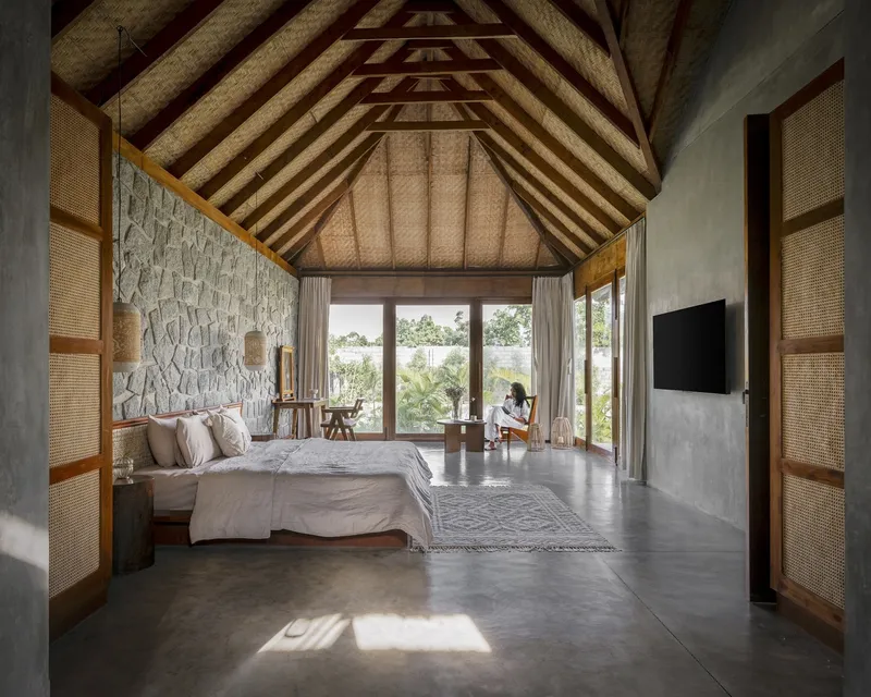 Bedroom with vaulted thatched roof and rattan paneled doors framing a figure seated by the window