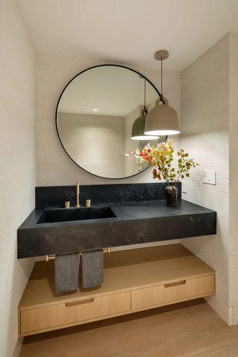 Floating black stone vanity with integrated sink beneath a large circular mirror and brass pendant light