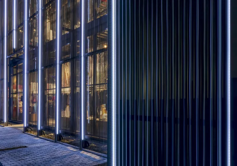 Illuminated vertical metal fins along a glazed facade and paved walkway at dusk