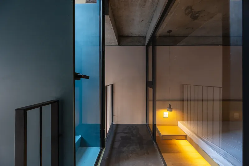 Narrow passage with exposed concrete ceiling looking through glazed doors to an illuminated room at dusk