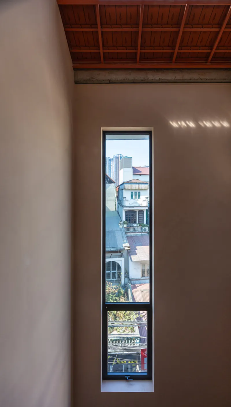 Tall narrow window framing neighboring rooftops beneath a timber coffered ceiling and concrete beam