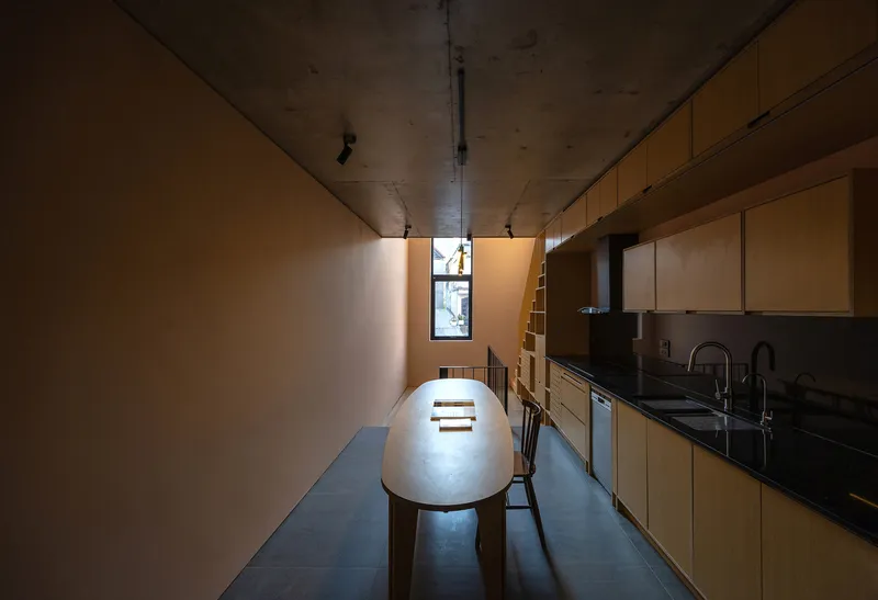 Galley kitchen with plywood cabinetry and concrete ceiling lit by narrow vertical window at the far end