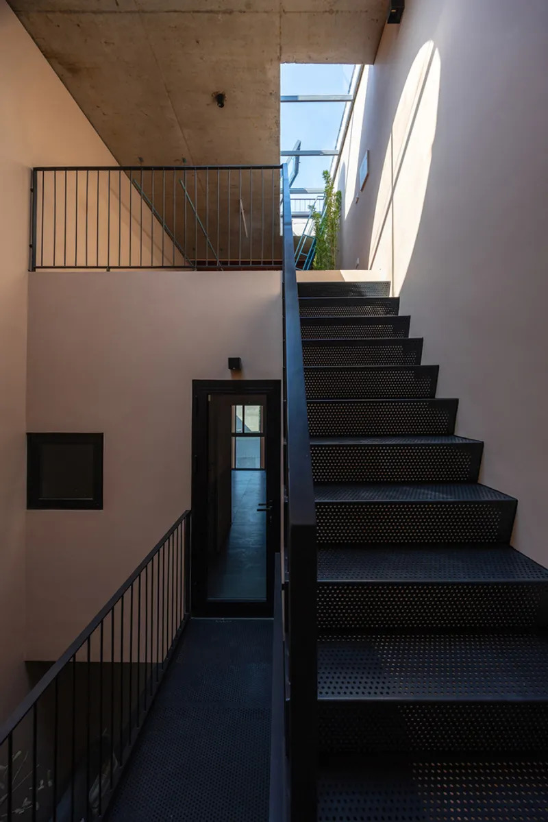 Black perforated steel staircase ascending through white walls with concrete ceiling and skylight above