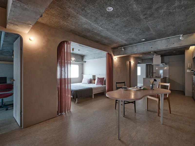Open-plan interior with curtained sleeping alcove, round dining table and exposed concrete ceiling at dusk