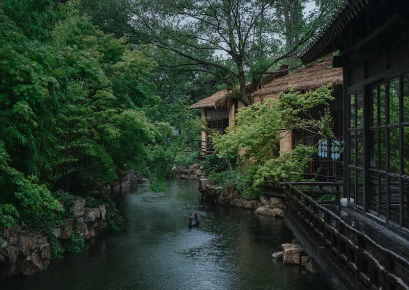 Thatched roof pavilions along a water channel edged with stacked stone and trees in overcast conditions