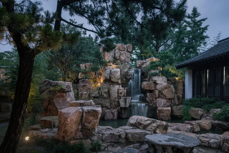 Stacked stone waterfall with integrated lighting among pine trees at dusk