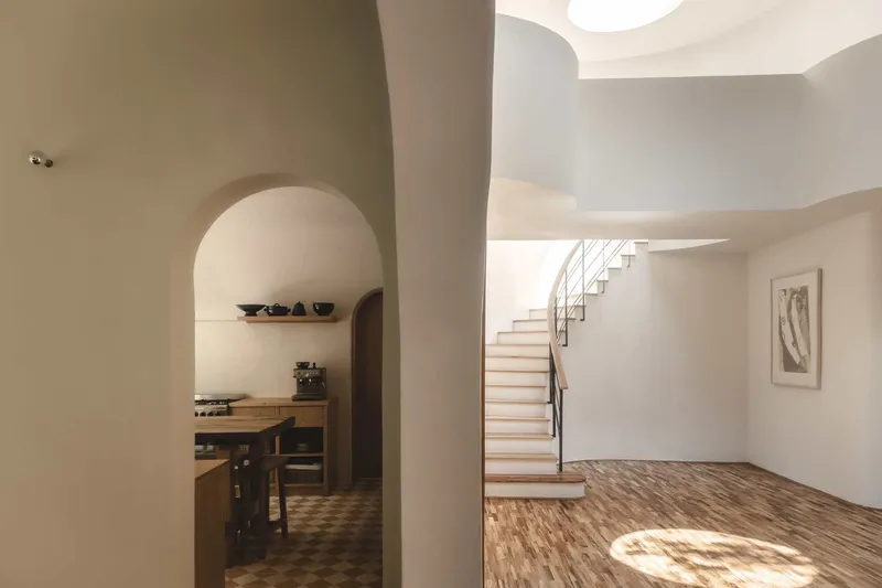Interior view through arched opening to kitchen nook and timber staircase with dappled sunlight on wood flooring
