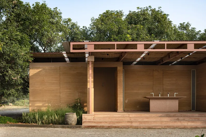 Open pavilion with rammed earth walls, exposed timber roof frame, and an outdoor sink counter flanked by grasses