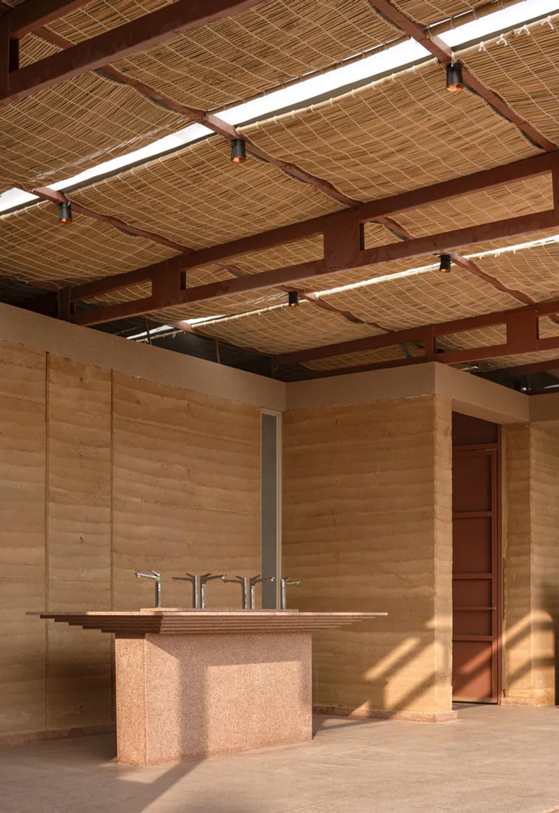 Interior showing woven bamboo ceiling panels, pink steel beams, and the rammed earth sink counter in sunlight