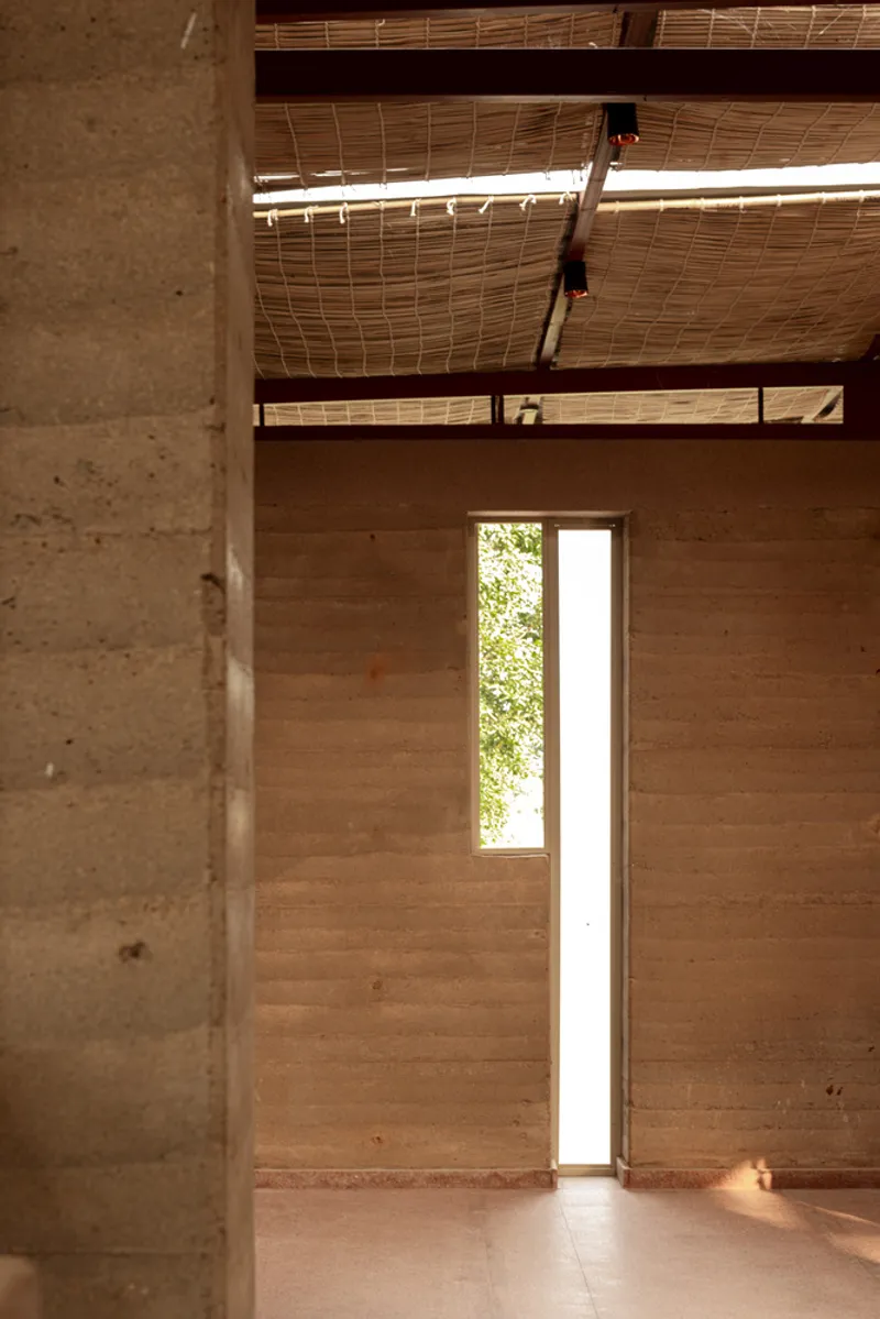 Double-height space with rammed earth walls, woven bamboo ceiling and narrow vertical window with sunlight