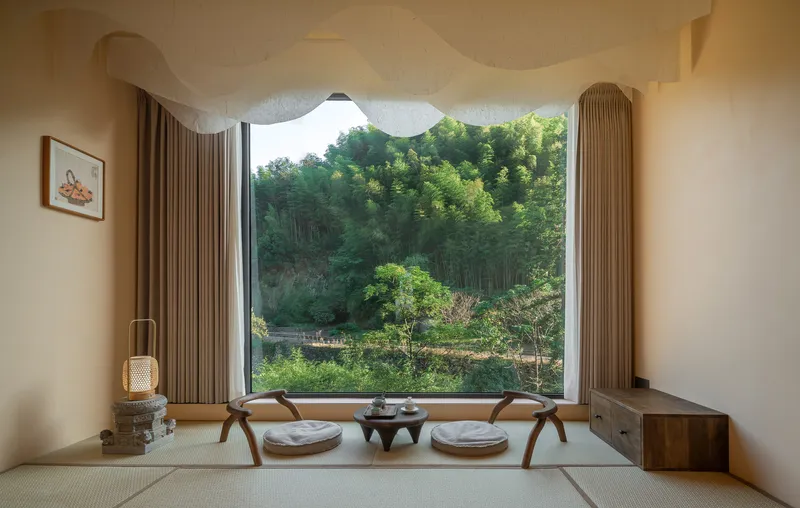 Tatami seating nook with low table and cushions facing a picture window overlooking terraced gardens and forest