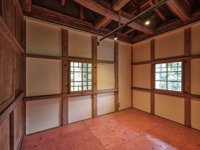 Room with exposed timber columns and beams, paper-paneled walls and two gridded windows admitting daylight