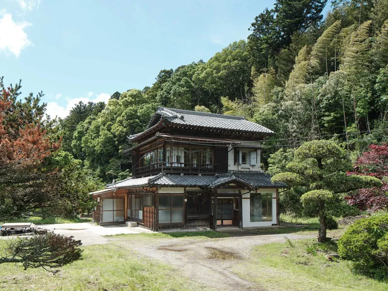 Two-story timber structure with tiled roof set against a forested hillside in summer