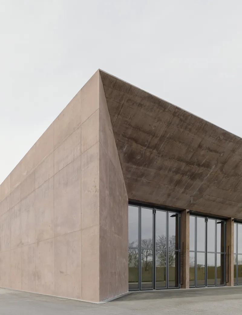 Corner view of the board-formed concrete volume with recessed glazed entrance under overcast sky