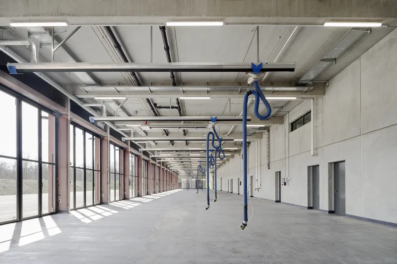 Interior workshop space with exposed concrete ceiling, blue chain hoists, and continuous glazing admitting daylight