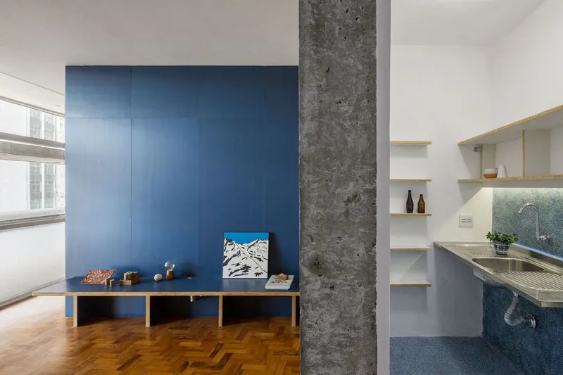 Living space with exposed concrete column, navy blue wall panel, open kitchen alcove, and herringbone parquet flooring