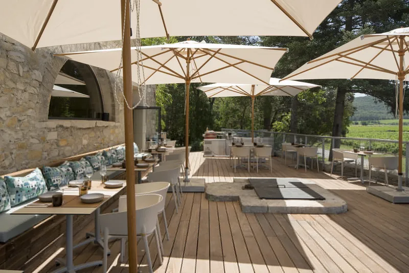 Dining terrace with timber decking, market umbrellas, stone archway, and vineyard views framed by trees