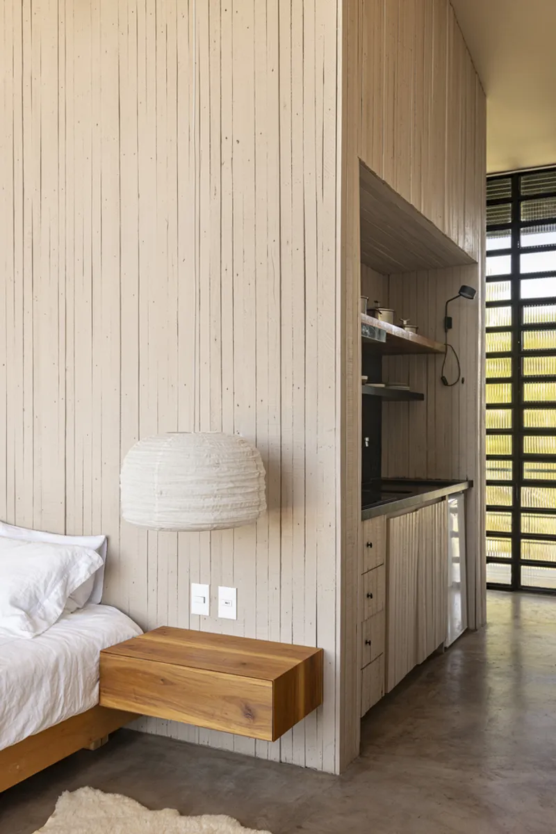 Bedroom with pale timber paneling, floating side table, and kitchenette niche beside translucent louvered window
