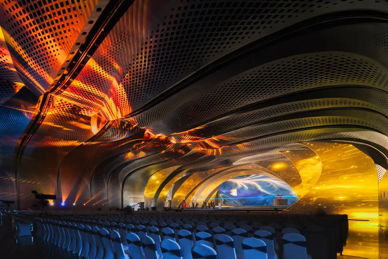 Perforated metal ceiling with orange and yellow LED lighting above rows of blue auditorium seating