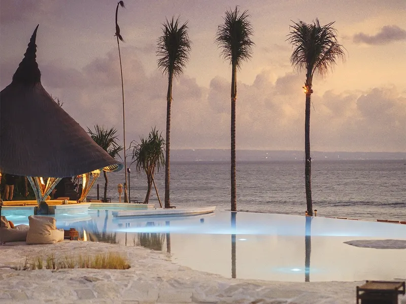 Infinity pool reflecting three tall palms and a thatched pavilion at dusk over the ocean