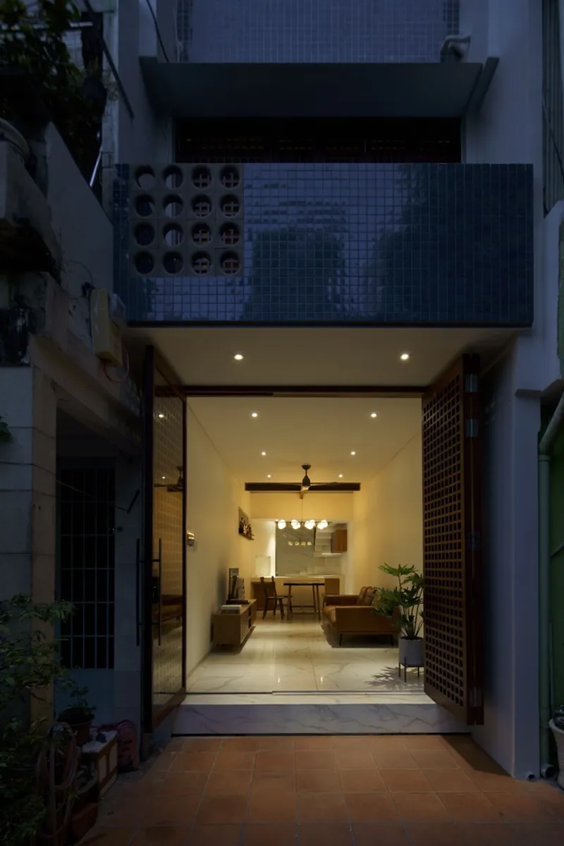 Street facade at dusk showing open glass wall revealing living room and tiled upper terrace with perforated screens