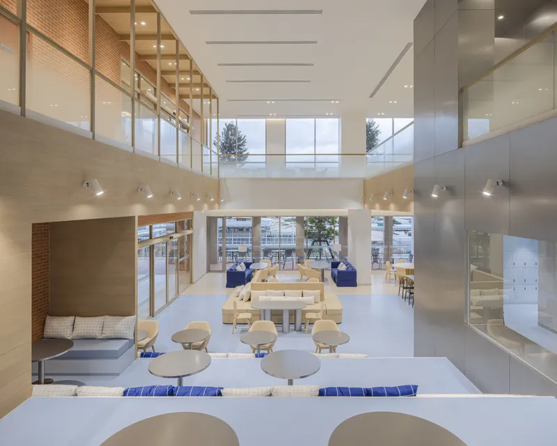 Two-story common area viewed from mezzanine with glass railings and blonde wood tables in sunlight