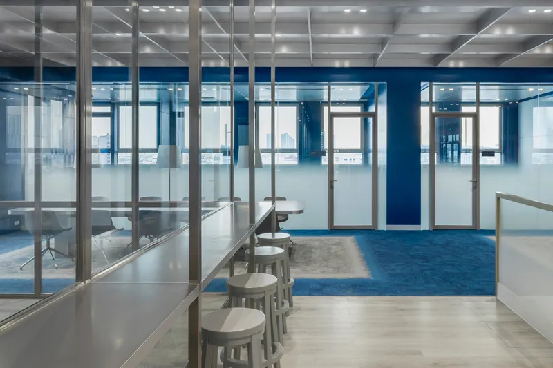 Corridor with blue carpet runner flanked by glass-walled rooms and white stools at low counters