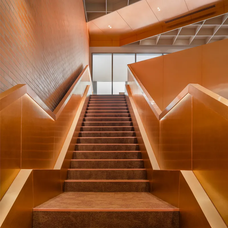 Central staircase with orange metal panel walls and dark terrazzo treads ascending toward diffused daylight
