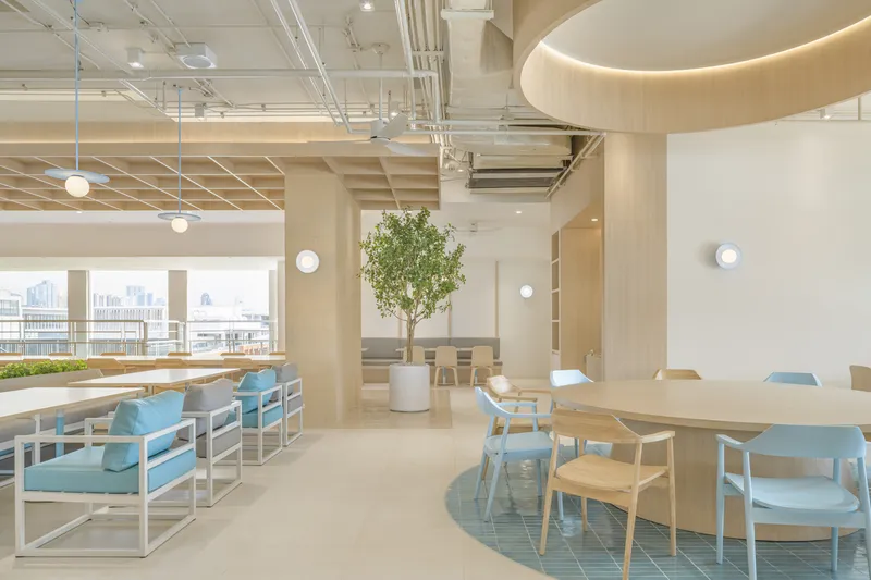 Open workspace with light wood tables, blue upholstered seating, exposed ceiling grid and potted tree