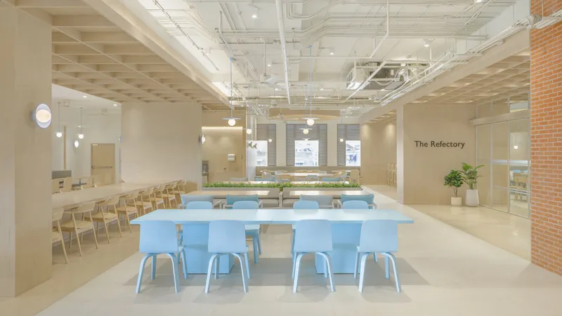 Long shared work tables with blue chairs beneath suspended pendant lights and exposed mechanical systems
