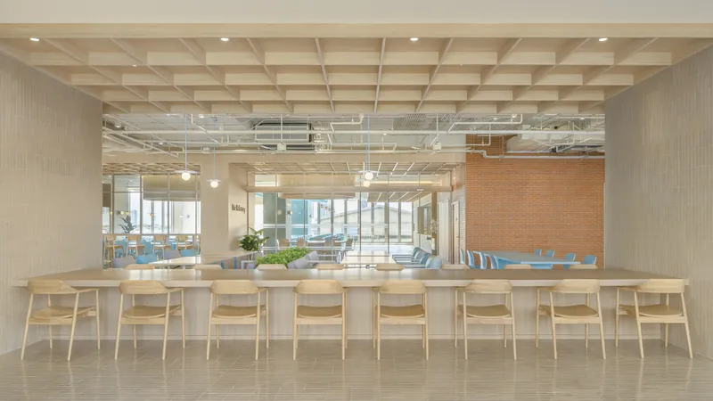 Continuous communal table with light wood chairs under grid ceiling with exposed conduit and brick wall