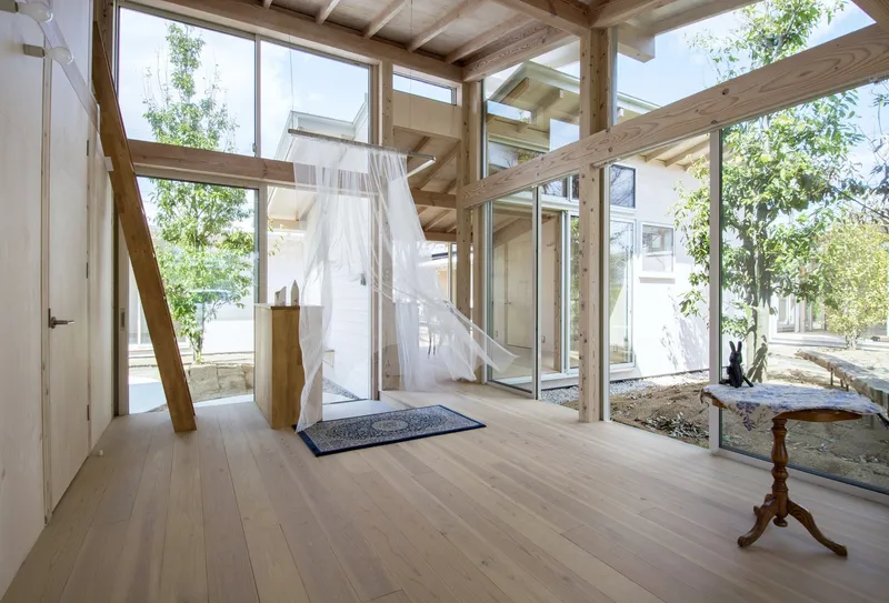 Bedroom with suspended white canopy bed beneath exposed timber structure and floor-to-ceiling windows overlooking garden