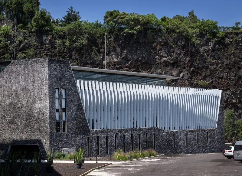 Cantilevered structure with vertical white fins against volcanic rock face under clear skies