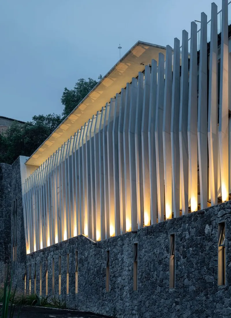 Illuminated vertical slat facade rising above a stone retaining wall at dusk