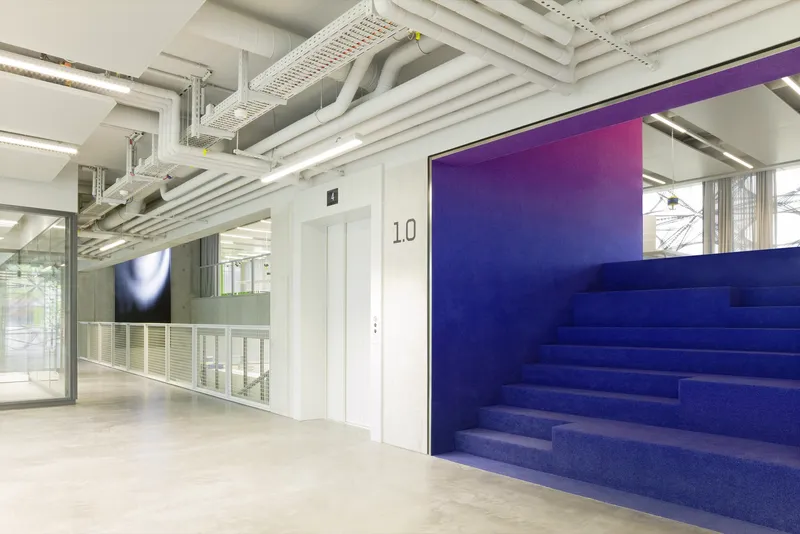 Blue carpeted stepped seating platform with gradient wall panel and exposed ceiling infrastructure