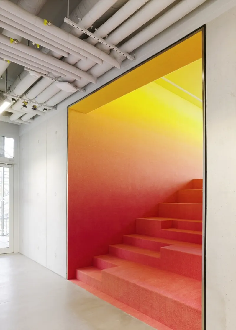 Interior staircase with gradient red-to-yellow wall and exposed mechanical ceiling ducts
