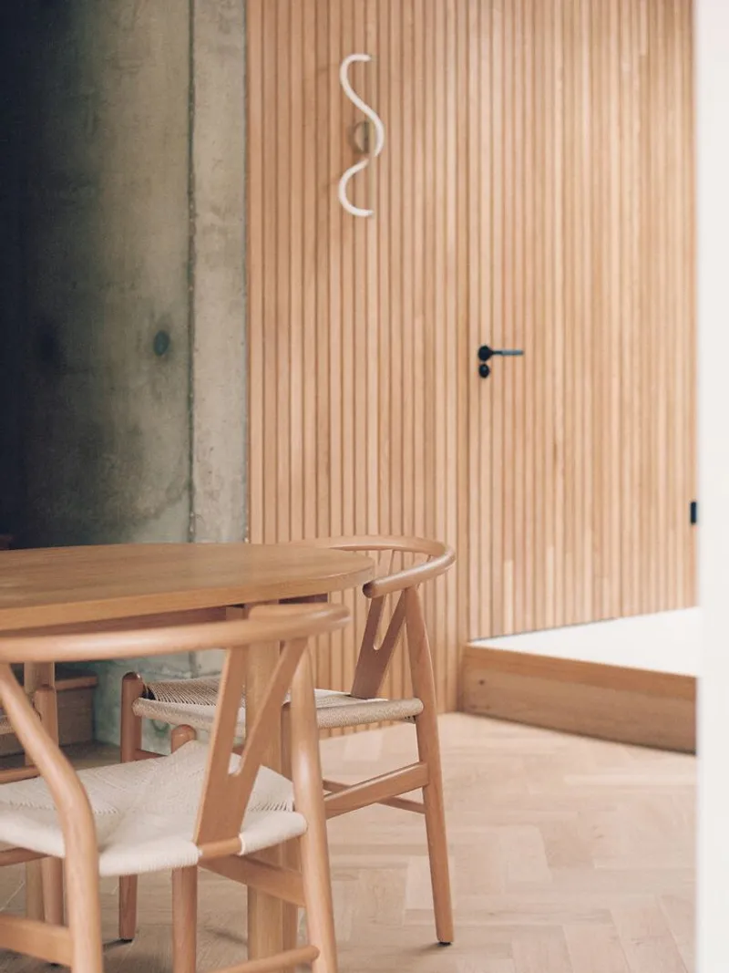 Dining table beside vertical timber slat wall with curved neon light fixture and exposed concrete column