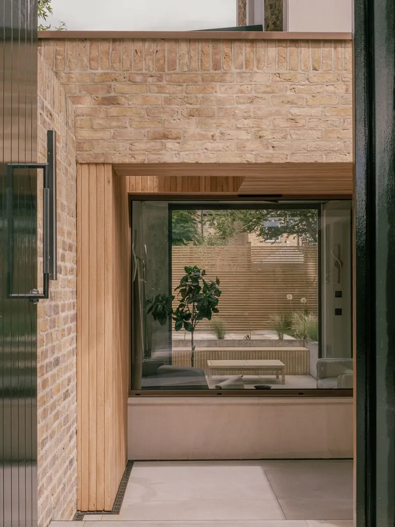 Courtyard view through timber-framed glazing toward an interior bench with potted fig tree