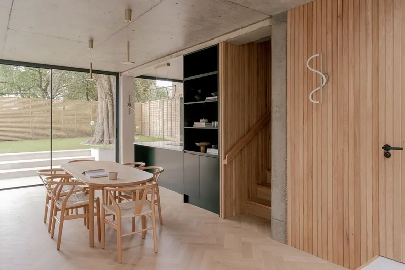 Dining area with timber-clad staircase and green shelving unit overlooking garden through glazed wall
